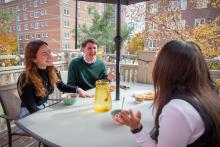 Three students at the Laurentian Leadership Centre sit around a table on an outdoor patio. The table holds a yellow pitcher, two bowls of food, and a glass with a drink. In the background are trees with autumn foliage and multi-story brick buildings.