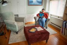 A student at the Laurentian Leadership Centre sits on a light-colored armchair in a bright room with wooden floors and white built-in cabinets, reading a book. A brown ottoman with papers and another book sits between two armchairs. A large window lets in natural light, and a bookshelf filled with books stands nearby.