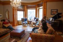 Five students at the Laurentian Leadership Centre sit in a living room with wooden floors and large windows, engaged in conversation. The room features a chandelier, a grand piano with sheet music, an air conditioning unit, artwork on the walls, a central rug, and an ottoman near the sofas.