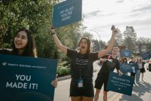 A group of university students wearing shirts with "Be transformed by the renewing of your mind. Romans 12:2" written on it walk outdoors holding welcome signs for Trinity Western University, celebrating new arrivals with messages like “YOU MADE IT!” and “SO GLAD YOU’RE HERE!”