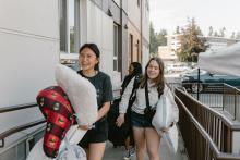Three young student women walk outside a building carrying pillows and personal belongings;