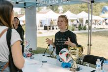 Two young women stand at a booth under a tent at an outdoor event, with a trophy, soccer ball, and papers on the table; other tents and attendees are visible in the background.