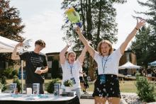 Two young women and a man stand outdoors near a table with brochures, a plant, and a candle; one young woman has her arms raised with a water gun in one hand, with trees and buildings in the background.