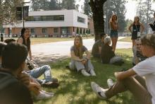 A group of university students sit and stand on a grassy area in front of a two-story modern building with brick and white paneling, surrounded by trees.