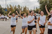 A group of university students run outdoors in white shirts, some cheering with big smiles and arms raised, with trees and a cloudy sky in the background.