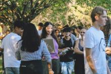 A group of young people gather and walk in a park-like outdoor setting, surrounded by trees and greenery; some hold drinks or yearbooks titled "PILLAR"