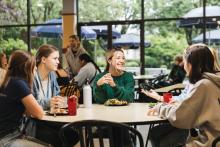 A group of young women sit around a table in a cafeteria, conversing over food and drinks, with large windows showing greenery and outdoor seating with umbrellas.