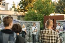 A group of musicians perform outdoors on a stage with instruments and microphones, while an audience watches; the background features a stone wall and trees.