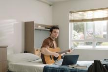 Student playing an acoustic guitar beside an open laptop in a residence room.