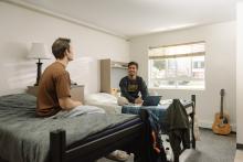 Two students talking in a furnished residence room with a laptop and guitar.