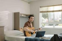 Student playing an acoustic guitar beside an open laptop in a residence room.