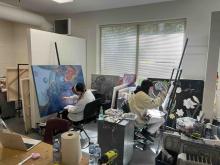 Two students painting large canvases in a studio surrounded by artworks and art supplies.