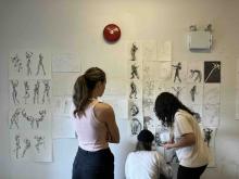 Three students reviewing figure drawings displayed across an art studio wall.