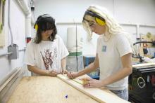 Two students wearing safety glasses and hearing protection measure and mark a wooden board in the SAMC woodworking shop.