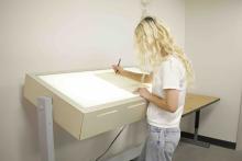 Student drawing on paper at an illuminated tracing table in a SAMC workspace.