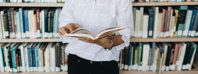 Student holding an open book in the library in front  of a shelf of books.