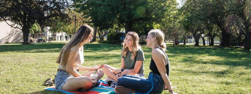 Three female students sitting on a blanket in the grass on campus talking.