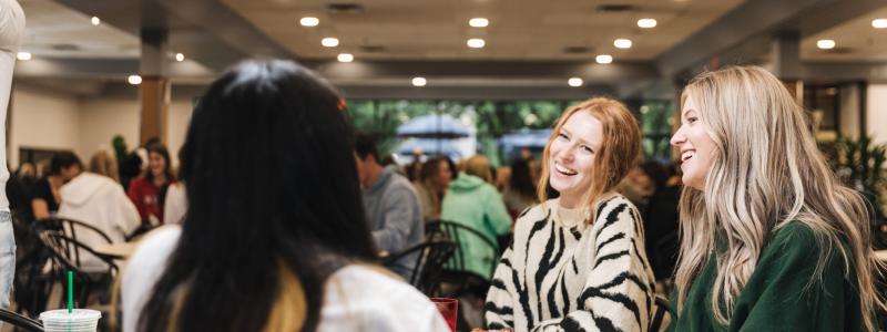 Three female students sitting in cafeteria eating, smiling, talking.