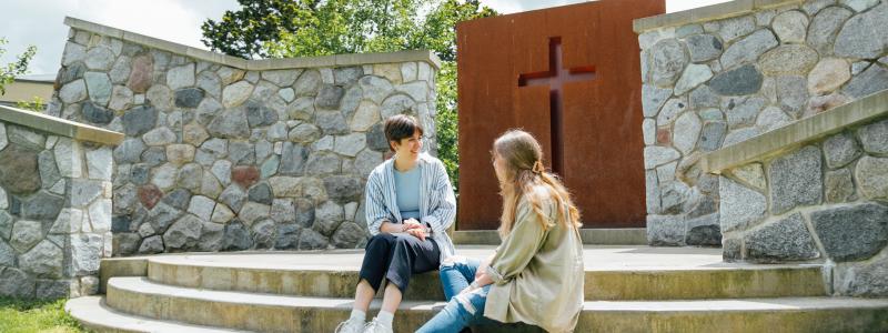 Two female students sitting on the steps of the Langley campus outdoor chapel talking. There is a cross in the background.