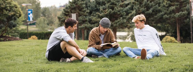 Three male students sitting on grass at the TWU Langley campus reading the bible and talking.