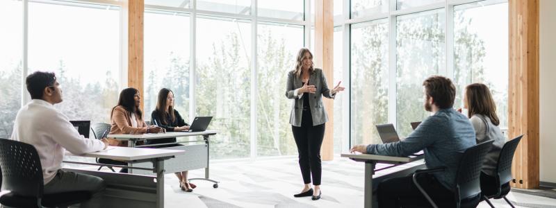 Teacher speaking to students sitting in a classroom with windows