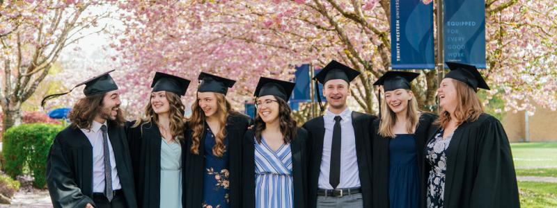 Seven students in graduation cap and gown smiling under the cherry blossoms with their arms around each other.