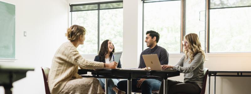 Four students sitting at a table studying on computers and laughing