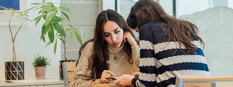 two students sitting at table and looking at phone together