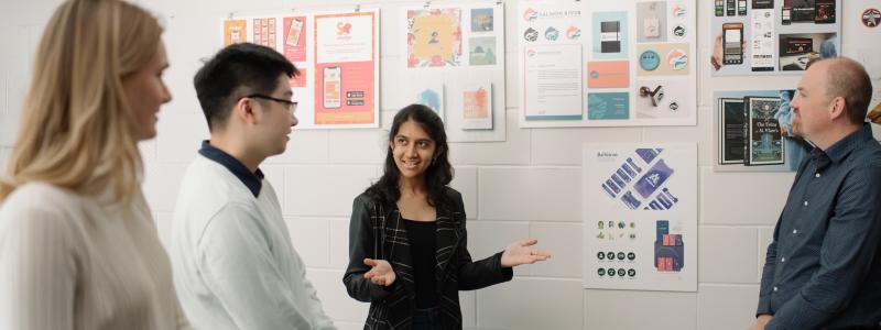 A group of people stand in front of a wall with posters displaying design projects. One person is speaking while others listen attentively.