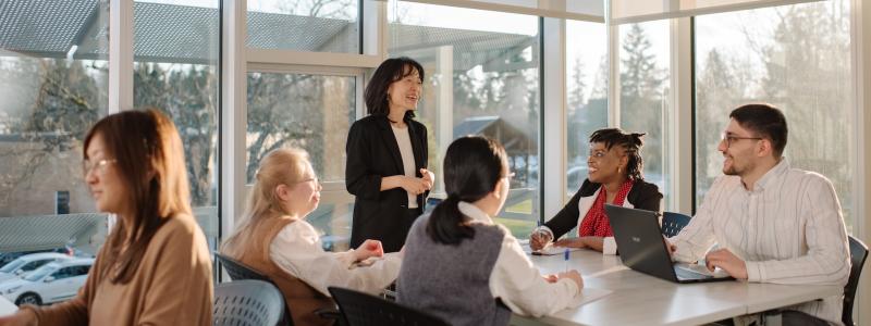 A group of people are seated around a table in a bright, modern office space with large windows. One person is standing and appears to be speaking or presenting to the group. The room has natural light coming through the windows, and there are trees visible outside.