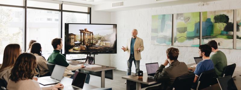 A group of university students seated with laptops in a classroom setting listen to a professor in front of a screen showing ancient ruins.