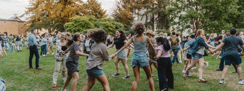 A group of people holding hands and dancing in a circle on a grassy field during an outdoor event.