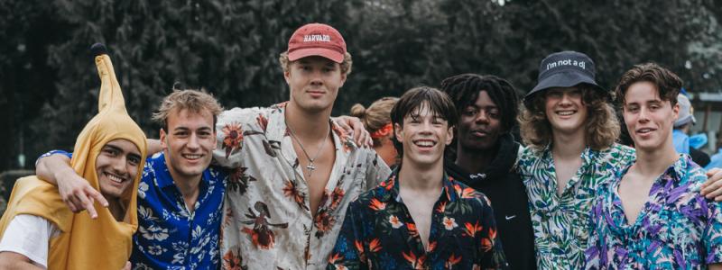 A group of seven people in tropical-themed outfits, including floral shirts and grass skirts, pose outdoors; one is dressed in a banana costume.