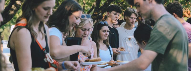 Young men and women standing in line at an outdoor buffet, serving themselves food in a park-like setting with trees in the background.