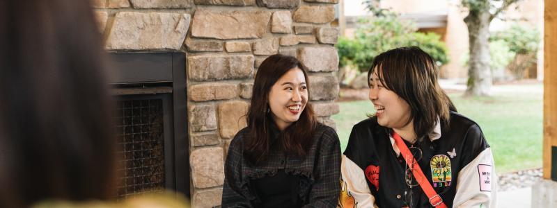 Two young women sit in front of a stone fireplace, smiling at each other. One holds a boba drink with a purple straw. A third person is partially visible in the foreground.