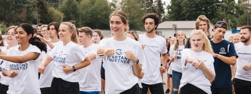 A group of young adults in white "SPARTANS TOGETHER" t-shirts participate in a synchronized outdoor activity, with trees and a building in the background.