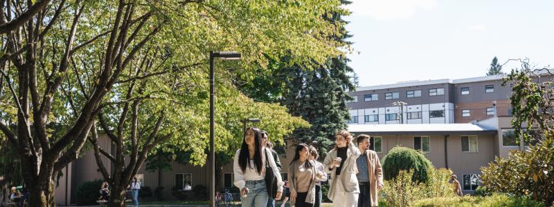 A group of people walk along a pathway in a campus-like setting, surrounded by trees, grass, and buildings.