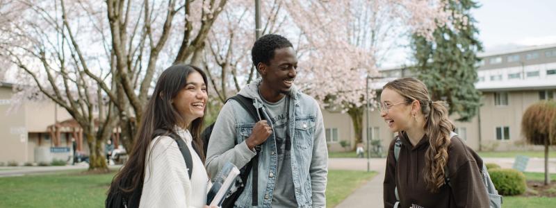Three students standing outdoors on a university campus, holding books and backpacks, with cherry blossom trees in the background.
