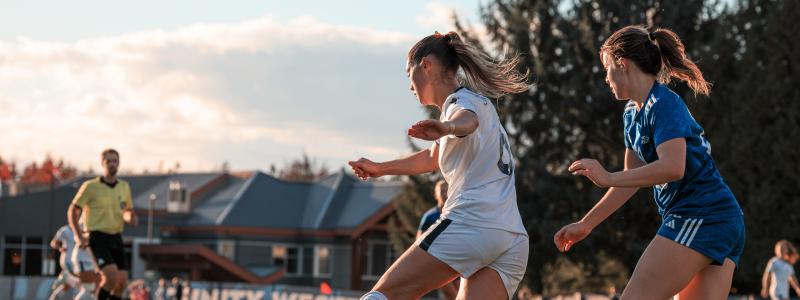 Two soccer players, one in white and one in blue, compete for the ball on an outdoor field, with a referee and teammates in the background. A "TRINITY WEST" banner is visible on the sideline.