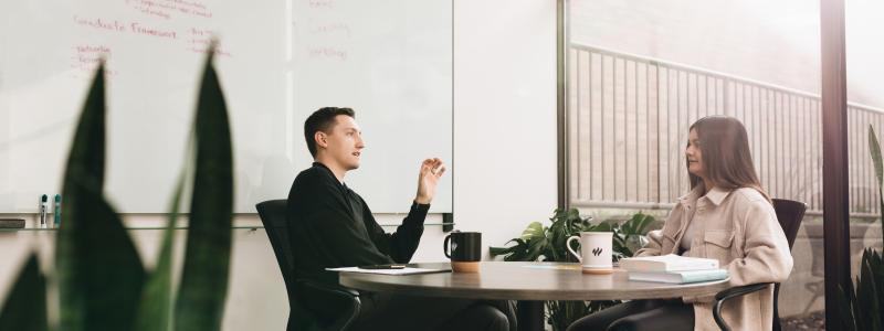 Two people sit at a round table in an office, talking, with a whiteboard behind them.