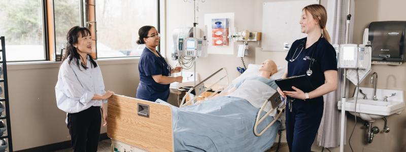 Three people stand around a patient in a hospital bed, surrounded by medical equipment and natural light from nearby windows.