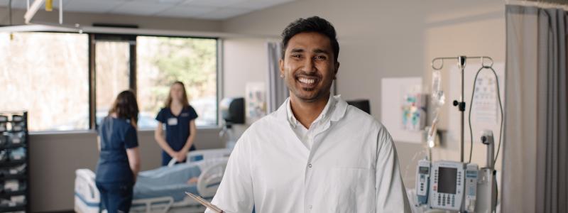 A person in a white lab coat holds a clipboard in a hospital room, while two others stand near a bed surrounded by medical equipment.