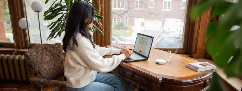 Person in a white sweater working on a laptop at a wooden desk near large windows with a city view.