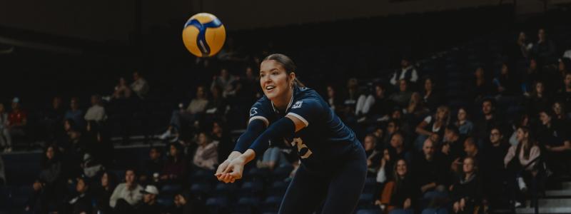 Volleyball player in a dark blue uniform prepares to receive the ball with arms extended.