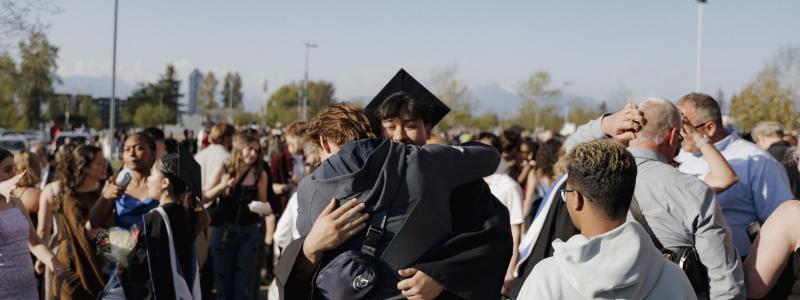 A person in a graduation cap and gown hugs someone during an outdoor ceremony, surrounded by people with flowers and gifts, with trees and mountains in the background.