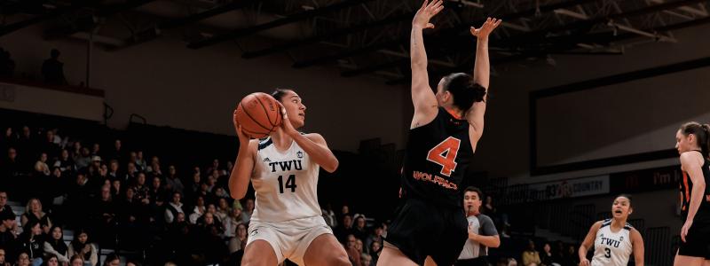 TWU basketball player in white jersey number 14 holds the ball while being closely defended by a player in black jersey number 4.