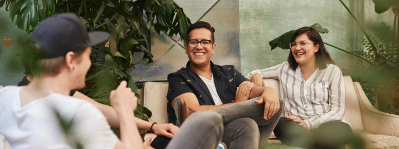 Three people laughing and chatting on a sofa surrounded by plants.