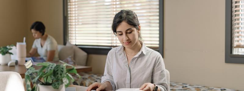 Woman reading a book at a table near a window with blinds.