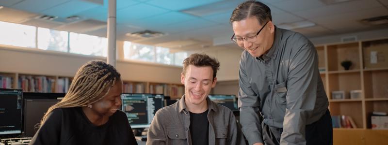 Three people smiling and collaborating in a library with a circuit board on the table.