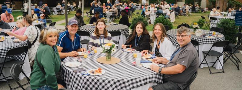 Group of people sitting at an outdoor table with checkered tablecloths, smiling.
